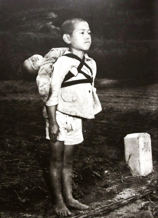 A boy in Nagasaki, is carrying his dead younger brother on his back and he's standing at a cremation pyre, trying to prepare himself to cremate his brother. (photo by Joe O'Donnell, 1945)